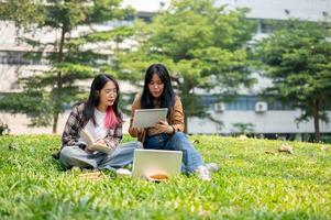 dos joven estudiantes son sentado en el césped en un parque, que se discute alguna cosa en un digital tableta. foto