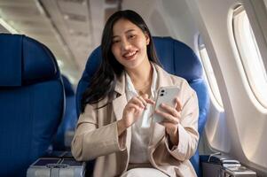 un atractivo asiático mujer de negocios es sentado a el ventana asiento con un teléfono inteligente en su mano. foto