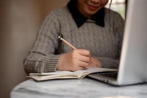 un joven asiático hembra estudiante es sentado adentro y escritura en su cuaderno con un lápiz. foto