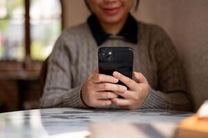 A cropped image of an Asian woman using her smartphone at a table indoors, and ordering goods online photo