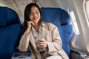 un atractivo asiático mujer de negocios es sentado a el ventana asiento con un teléfono inteligente en su mano. foto