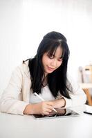 A young Asian woman using her digital tablet with a stylus pen at a table indoors. photo