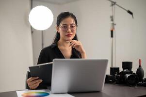 un atractivo asiático hembra fotógrafo es mirando a el cámara mientras sentado a un escritorio en un estudio foto