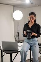 profesional asiático hembra fotógrafo es enfoque en ajustando su dslr cámara, trabajando en un estudio foto