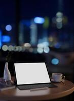 un pantalla blanca digital tableta Bosquejo en un escritorio en un moderno oficina a noche con ciudad noche vista. foto