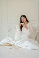 A beautiful Asian woman in white long dress pajamas is having breakfast on bed, sipping tea. photo