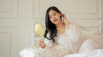 An attractive Asian woman in long dress pajamas is looking in the mirror while she is on her bed. photo