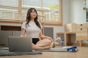un calma, relajado joven asiático mujer en ropa de deporte es meditando en un yoga estera en el vivo habitación. foto