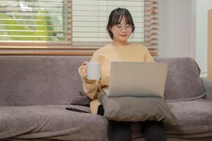 An attractive Asian woman is working remotely at home, sipping coffee, and working on her laptop. photo