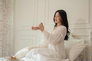 An attractive Asian woman in long dress pajamas is applying perfume after waking up on her bed. photo