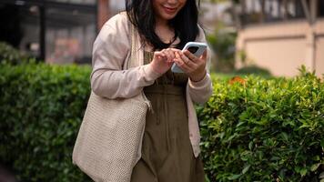 A happy Asian woman in casual clothes using her smartphone while walking in the city on a bright day photo