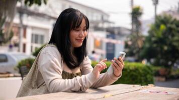 un atractivo, contento asiático mujer sentado a un mesa al aire libre en el ciudad y utilizando su teléfono inteligente foto