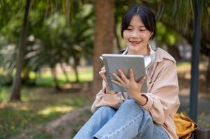 A positive, smiling young Asian woman is using her digital tablet on a bench in a park. photo