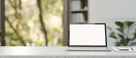 A white-screen laptop mockup, a clipboard paper, and a smartphone on a table in a modern office. photo