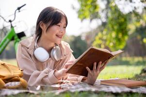 A happy Asian woman is enjoying reading a book on a picnic cloth, while chilling in a green park. photo