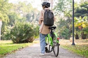 A young Asian woman is walking with her bicycle in a green park. back view image photo