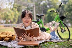 un contento asiático mujer es disfrutando leyendo un libro en un picnic paño, mientras Relajado en un verde parque. foto