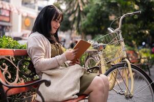 A positive Asian woman is reading a book while sitting on a bench outdoors in the city with her bike photo