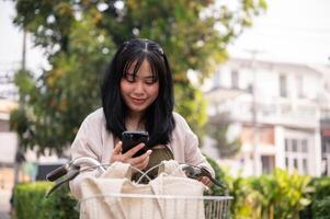 un hermoso, contento joven asiático mujer en un linda vestir es utilizando su teléfono inteligente en su bicicleta en el ciudad foto