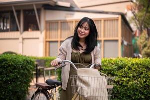 A beautiful, positive Asian woman in a cute dress is walking and pushing her bicycle in the city. photo