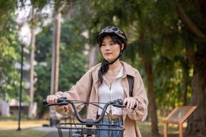 A young Asian female college student wearing a bike helmet is pushing her bicycle in a green park. photo