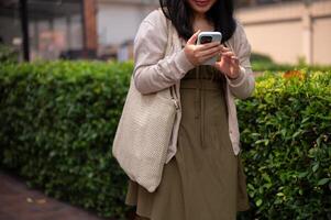 A happy Asian woman in casual clothes using her smartphone while walking in the city on a bright day photo