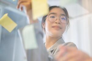 un creativo asiático mujer de negocios leyendo algunos ideas en pegajoso notas mientras lluvia de ideas con su equipo. foto