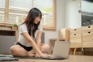 un encantador joven asiático mujer sentado en un yoga estera y utilizando su ordenador portátil computadora a hogar. foto