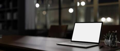 A modern dark office at night features a white-screen laptop computer mockup on a table. photo