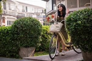 A friendly Asian woman waves her hand to say hi to her friend while pushing her bike in the city. photo