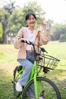 A cheerful and smiling young Asian woman is riding her bike in the green park on a sunny day. photo