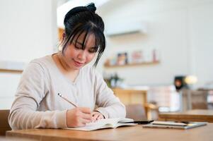 A positive Asian woman is writing in a book, working on her tasks while sitting in a coffee shop. photo