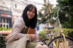 An attractive, happy young Asian woman is sitting on a bench outdoors in the city with her bike. photo