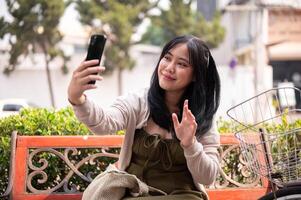 A positive Asian woman is talking on a call with someone while relaxing on a bench outdoors. photo