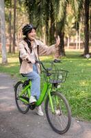 un contento joven asiático hembra Universidad estudiante es disfrutando montando su bicicleta en un verde parque. foto