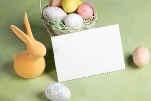 White blank note on pastel yellow green backdrop surrounded by color Easter eggs and yellow bunny. photo