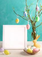 Easter card. White frame with copy space, twigs with spring leaves, color eggs on them on turquoise. photo