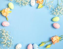 Frame of decorative Easter eggs and delicate white and peach flowers on blue. Top view. Copy space. photo