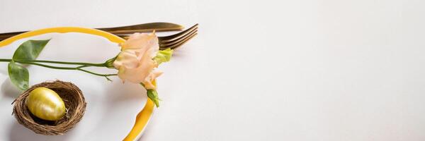 Easter banner. Gold egg in nest, Eustoma flower on white yellow plates, gold cutlery on white pink. photo