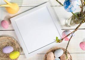 Empty white frame, color Easter eggs, yellow bunny, spring twigs on white wooden table. Top view. photo