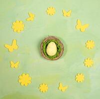 Easter egg in nest inside of round frame of yellow flowers and butterflies on green yellow. Top view photo