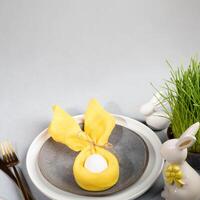 Plates, white Easter egg in yellow napkin, grass, ceramic bunnies, gold cutlery on gray. Copy space. photo