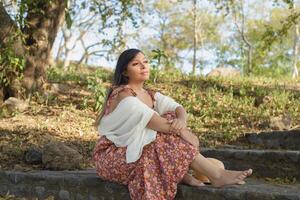 mujer vistiendo sombrero y vestir con flores, descalzo sentado en el escalera de un público parque. soleado verano día. foto