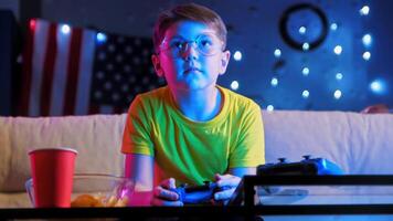 A little boy with glasses is enthusiastically playing a game on a game console, using a gamepad, against a backdrop of a garland and a US flag, under the bright glow of neon lights. Mid shot. 4k video