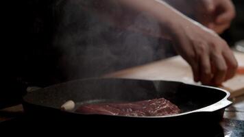 Cook adds pieces of garlic and carrots to the beef as it roasts in a hot frying pan, with vegetable oil splashing on a dark background. Close up. Slow motion. 4k video