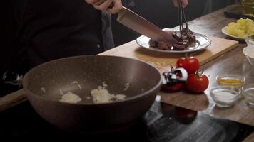 een koken in een zwart uniform is voorbereidingen treffen een schotel in de keuken van een restaurant. ze zijn snijden sappig rundvlees Aan een bord, en in de voorgrond, uien zijn wezen gebakken in een keramisch frituren pan. dichtbij omhoog. langzaam beweging. 4k video