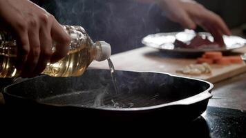 Pour des hommes mains verser légume pétrole sur une fumant, noir céramique friture la poêle dans une restaurant cuisine, où le le fourneau est alimenté par induction. proche en haut. lent mouvement. 4k video