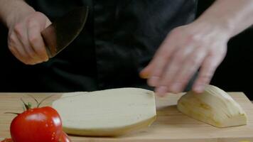 kochen, gekleidet im ein schwarz Uniform gegen ein schwarz abstrakt Hintergrund, ist Schneiden Zucchini in groß Scheiben auf ein hölzern Schneiden Tafel. Kamera dann zieht zurück, Aufschlussreich andere Gemüse Das wurden vorher versteckt. schließen hoch. schleppend Bewegung. 4k video