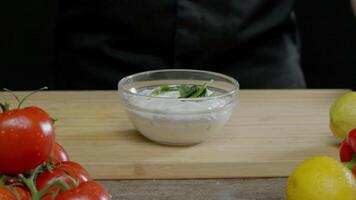 uma cozinhar dentro uma Preto uniforme, em pé contra uma Preto pano de fundo, é jogando peças do verde para dentro uma branco molho em uma mesa cercado de colorida legumes e frutas dentro a cozinha do a de luxo restaurante. fechar acima. lento movimento. 4k video