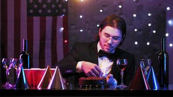 A young, lonely long-haired brunette birthday boy flows candles into a birthday cake. He is sitting at a festive table in a suit and glasses. Mid shot. 4k video